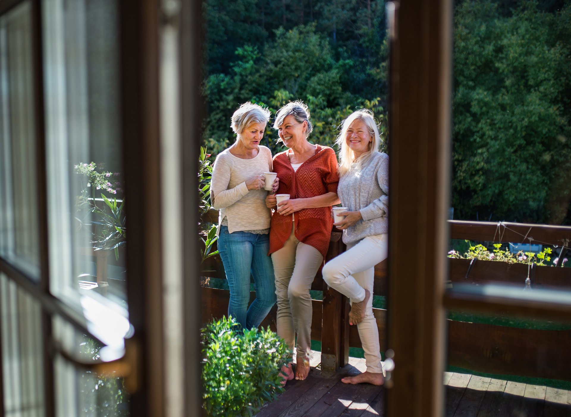 Drei Frauen stehen lächelnd und mit Tassen in der Hand auf einer Holzterrasse, die im Hintergrund begrünt ist.