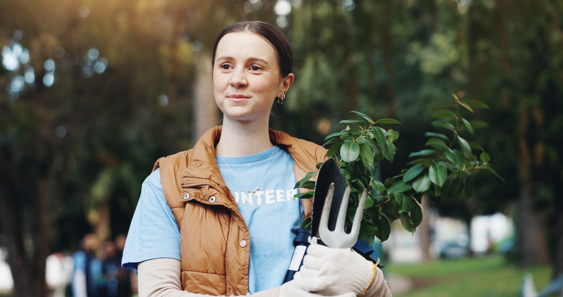 Eine Frau mit einer Pflanze und Gartengeräten in der Hand, die ein blaues „Volunteer“-Shirt und eine braune Weste trägt, steht im Freien.