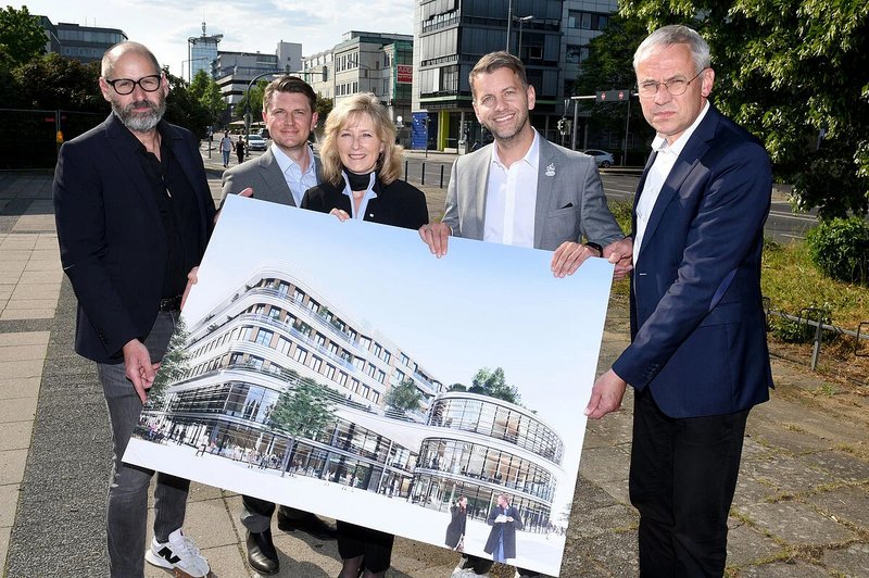 Fünf Personen halten ein großes architektonisches Rendering eines modernen mehrstöckigen Gebäudes, das im Freien auf einer gepflasterten Fläche mit Bäumen und Gebäuden im Hintergrund steht.